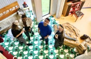 Photo from bird view, looking down at four people, one of them looking up to the camera. The floor they are standing on is colourful, with green and white patterns and there is colourful furniture seen on the background.