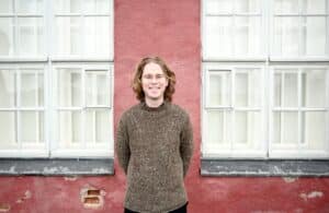 White man with curly blonde hair smiling in front of a pink building with white windows.